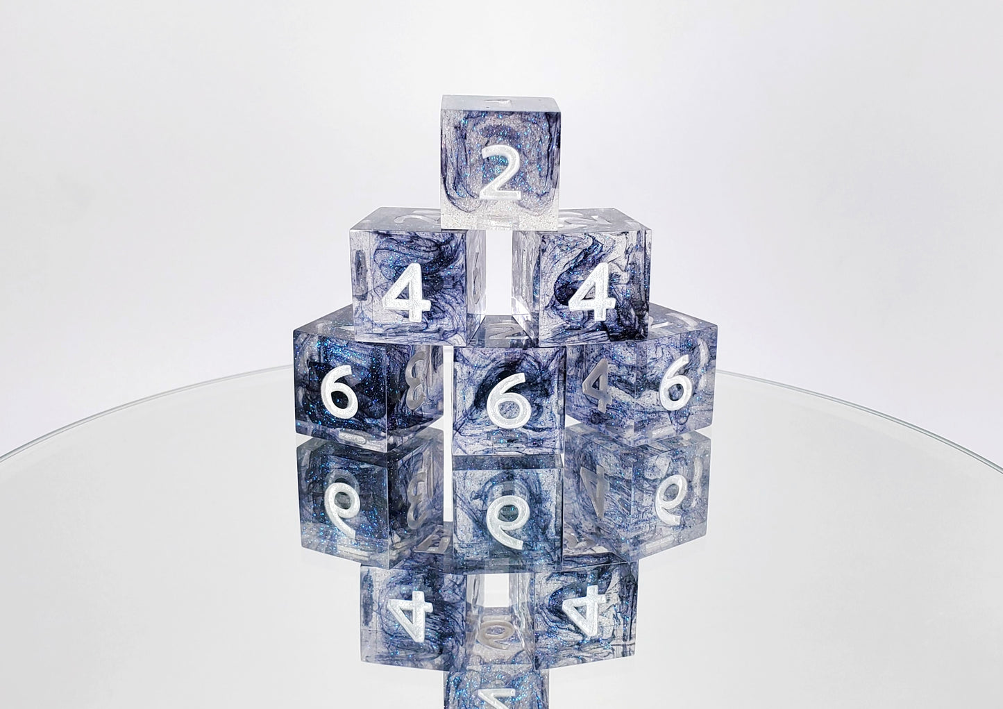A stack of 6 d6s. The dice are clear resin with blue/pink colorshift sparkles and wisps of black with pearl white numbers.