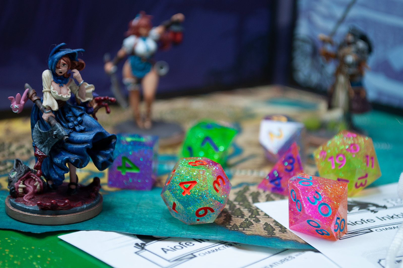 A 7 piece glitter dice set on top of a coastal map surrounded by 3 painted character miniatures. The dice are each 2 to 3 glitter colors, covering the whole rainbow across the entire set, with varying number colors to compliment each die.