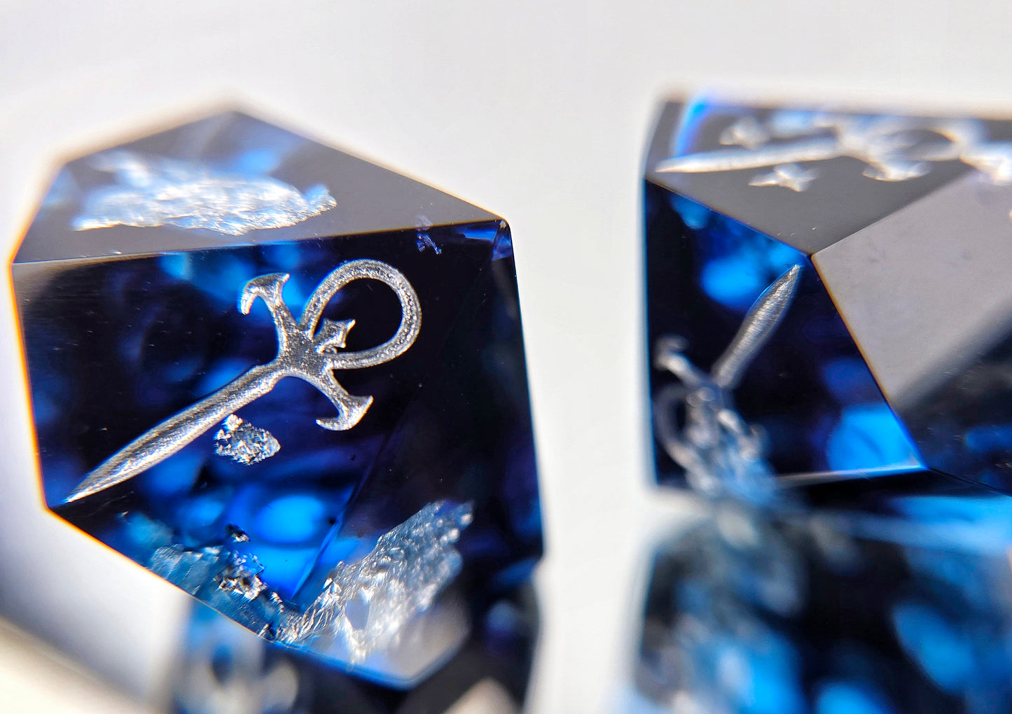 A closeup of a Vampire the Masquerade Hunger style die focusing on the plain ankh symbol. The dice are transparent blue with black ink and silver foil inside. The symbols are inked in silver.