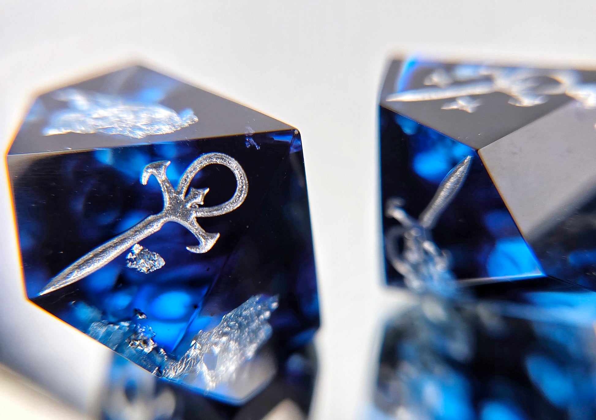 A closeup of a Vampire the Masquerade Hunger style die focusing on the plain ankh symbol. The dice are transparent blue with black ink and silver foil inside. The symbols are inked in silver.