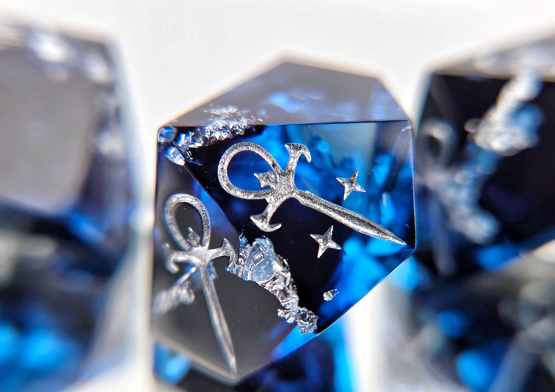 A closeup of a Vampire the Masquerade Hunger style die focusing on the star ankh symbol. The dice are transparent blue with black ink and silver foil inside. The symbols are inked in silver.