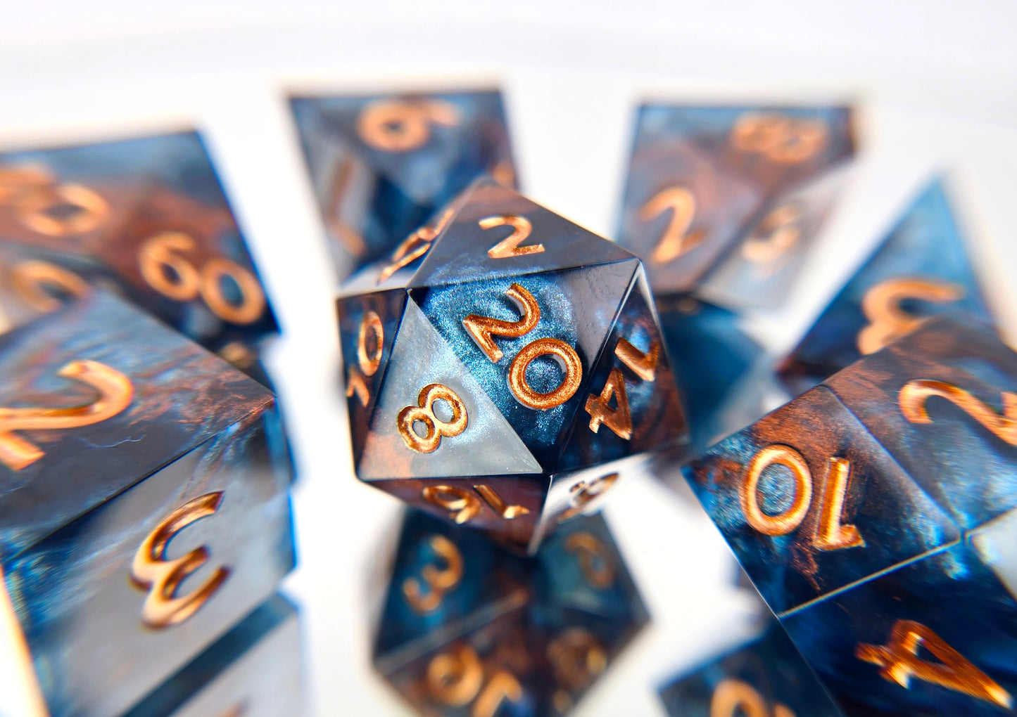 A full 7 piece polyhedral ttrpg dice set. The dice are transparent dark blue with bloops of shimmery silver inside and streaks of bronze coloring across the surface and warm copper numbering.