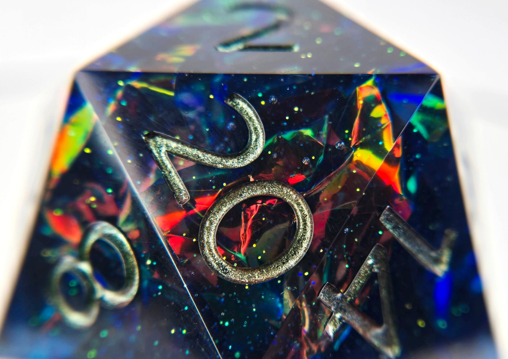 A closeup of an oversized d20, with the interior in focus. The die is a transparent dark blue resin with super fine green glitter sparkles and intense red to orange to yellow to green colorshift mylar foil crinkles inside. The numbers are inked a greenish bronze.