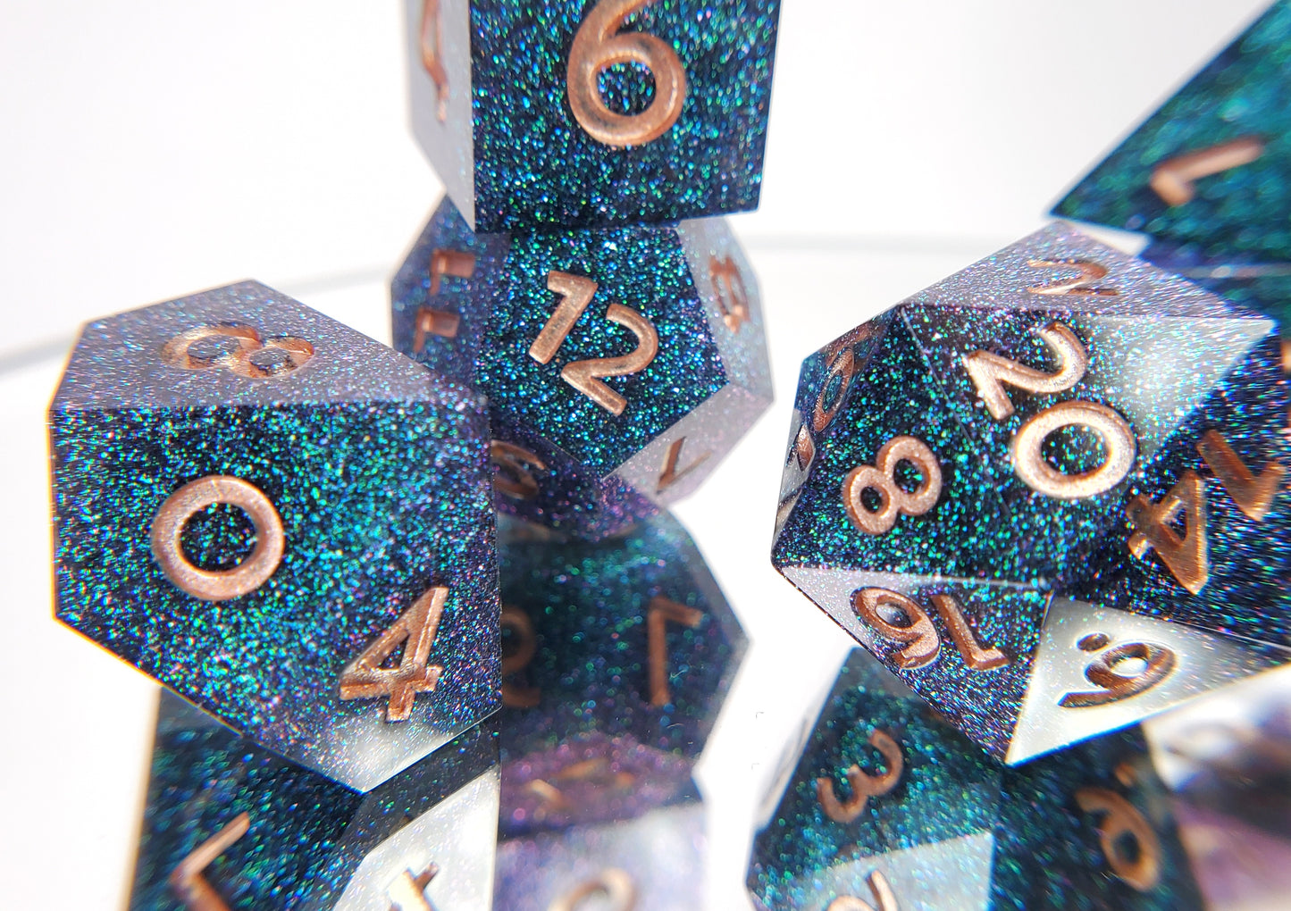 A closeup of a d10from a ttrpg set with the rest of the dice out of focus around and behind it. The dice are densely packed blue and green colorshift glitter with rose gold inking for the symbols.