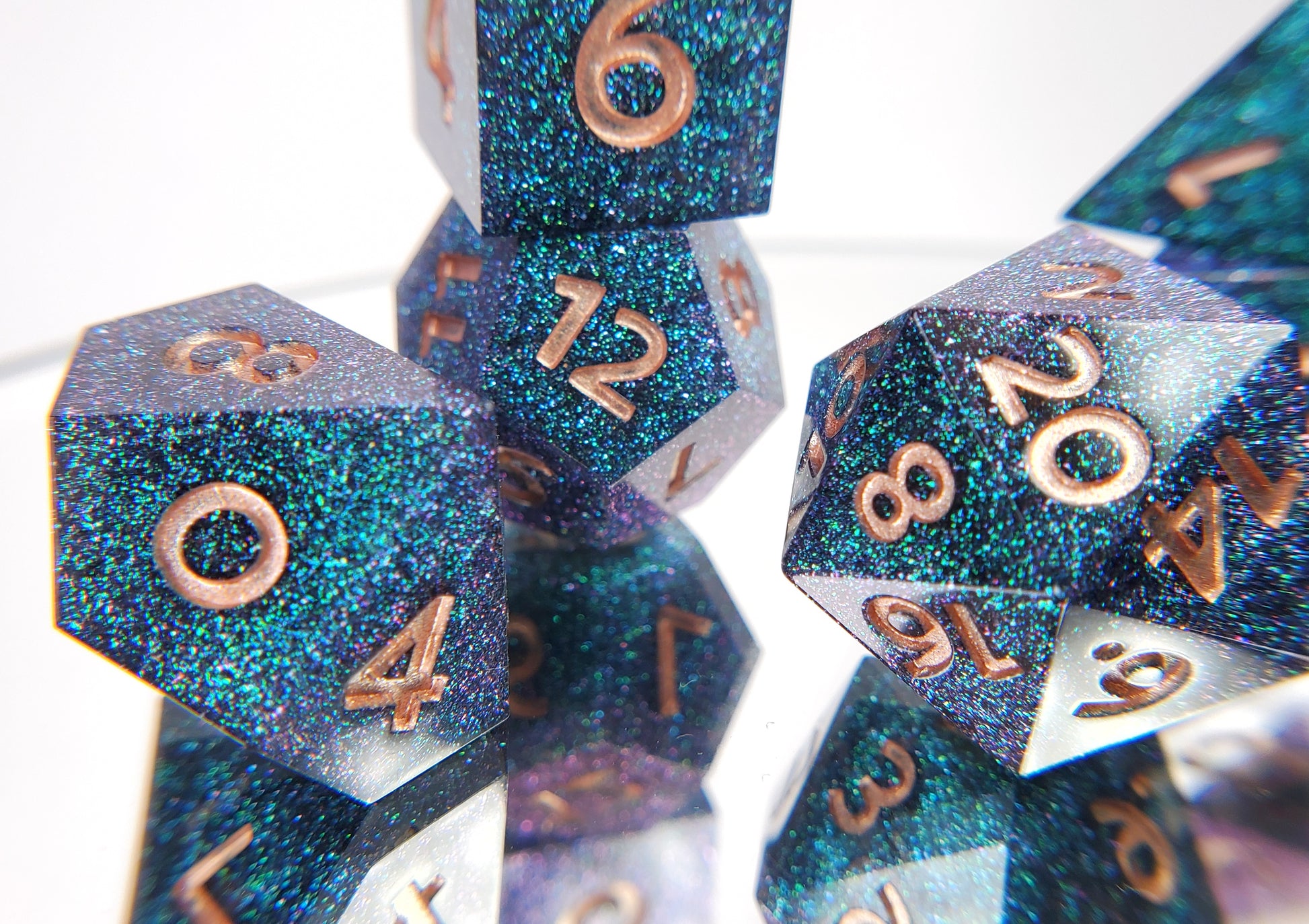 A closeup of a d10from a ttrpg set with the rest of the dice out of focus around and behind it. The dice are densely packed blue and green colorshift glitter with rose gold inking for the symbols.