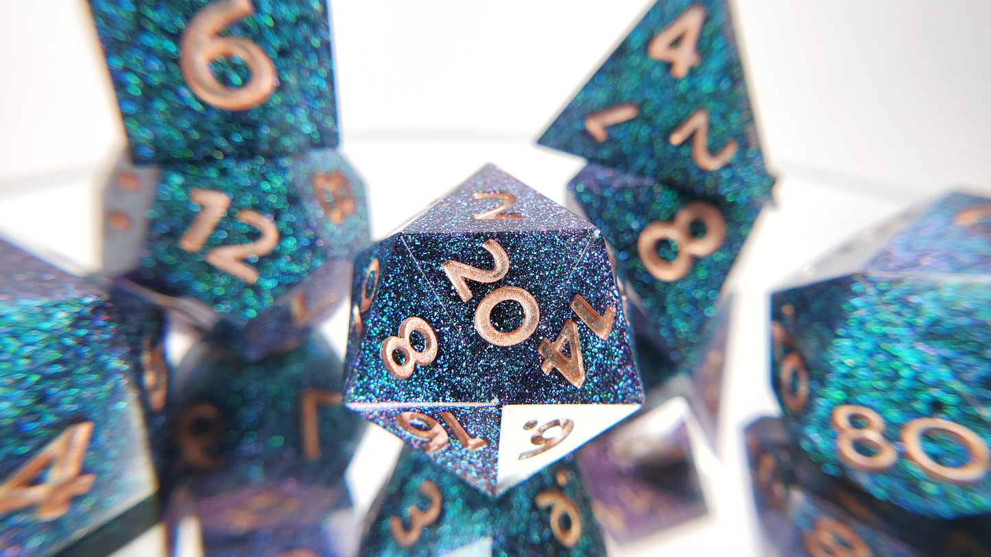 A closeup of a d20 from a ttrpg set with the rest of the dice out of focus around and behind it. The dice are densely packed blue and green colorshift glitter with rose gold inking for the symbols.