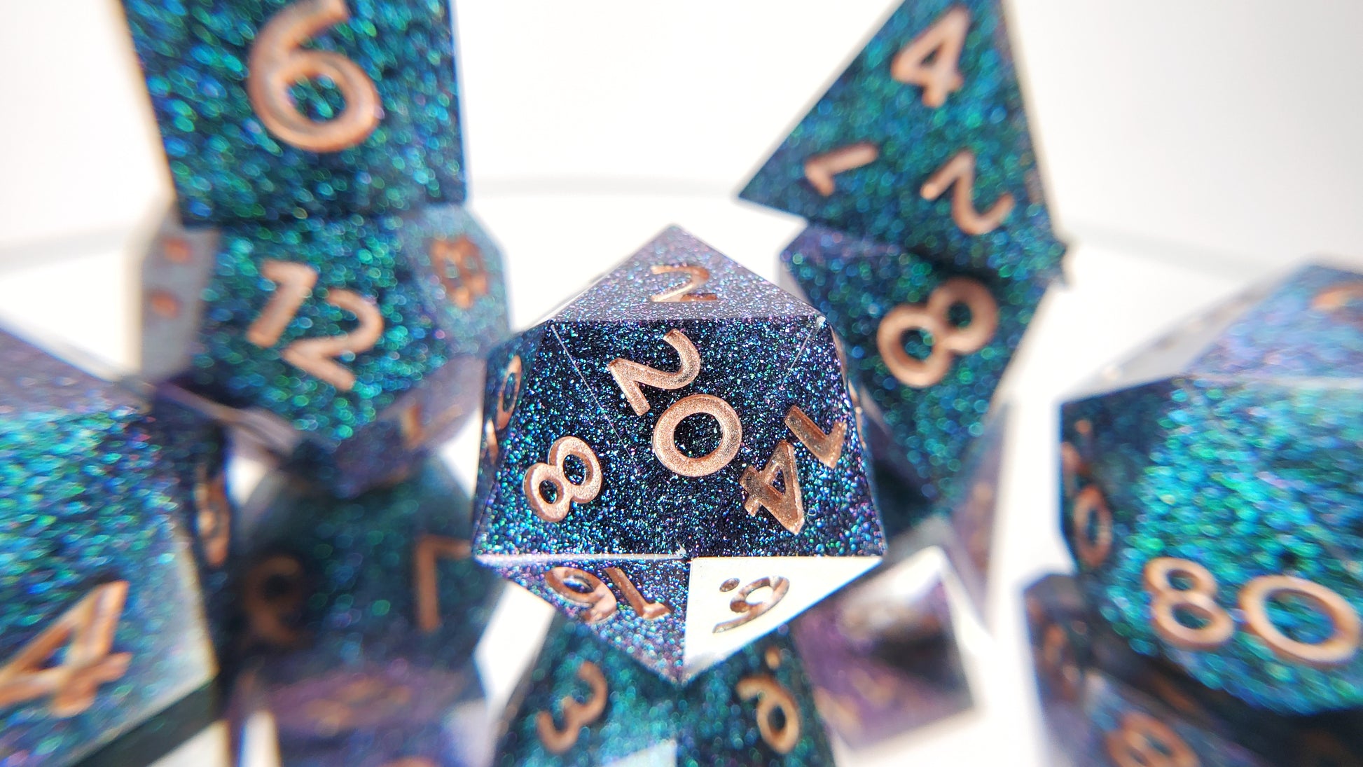 A closeup of a d20 from a ttrpg set with the rest of the dice out of focus around and behind it. The dice are densely packed blue and green colorshift glitter with rose gold inking for the symbols.