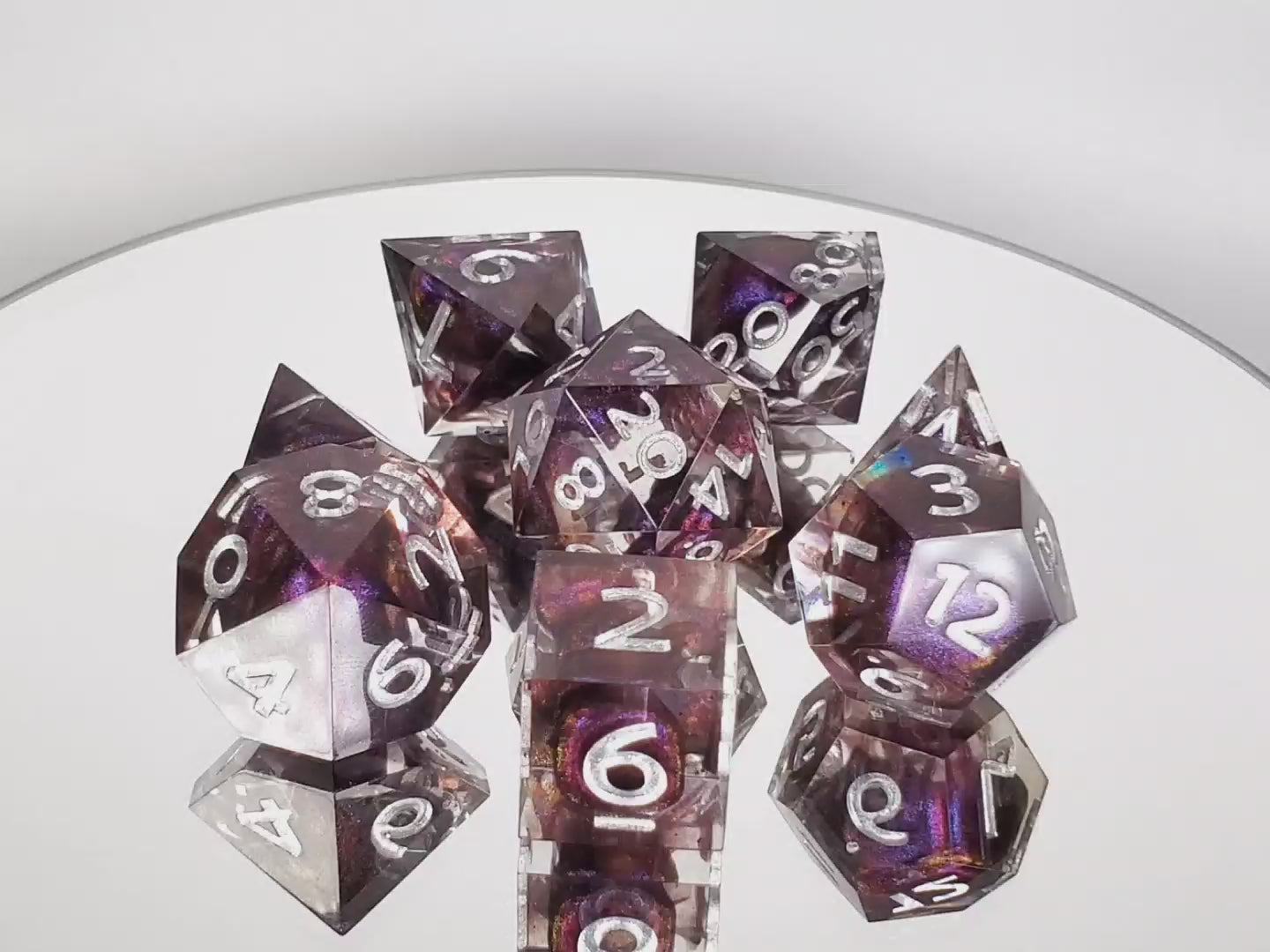 Polyhedral dice set of clear resin with dark purple colorshifting shimmer bloops and silver numbering on a rotating mirror platform.