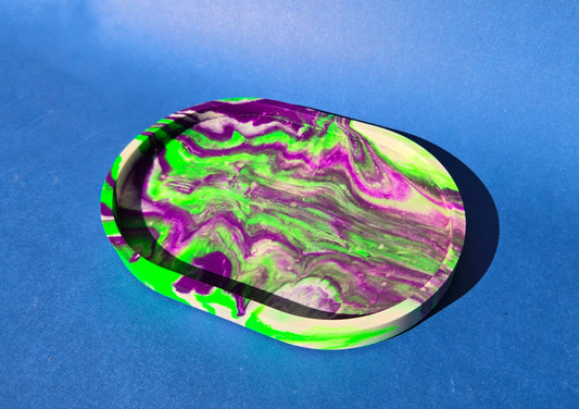 A jesmonite oval trinket tray or catch-all tray with bright green and purple swirls across the natural off-white surface. The tray is against a blue background.