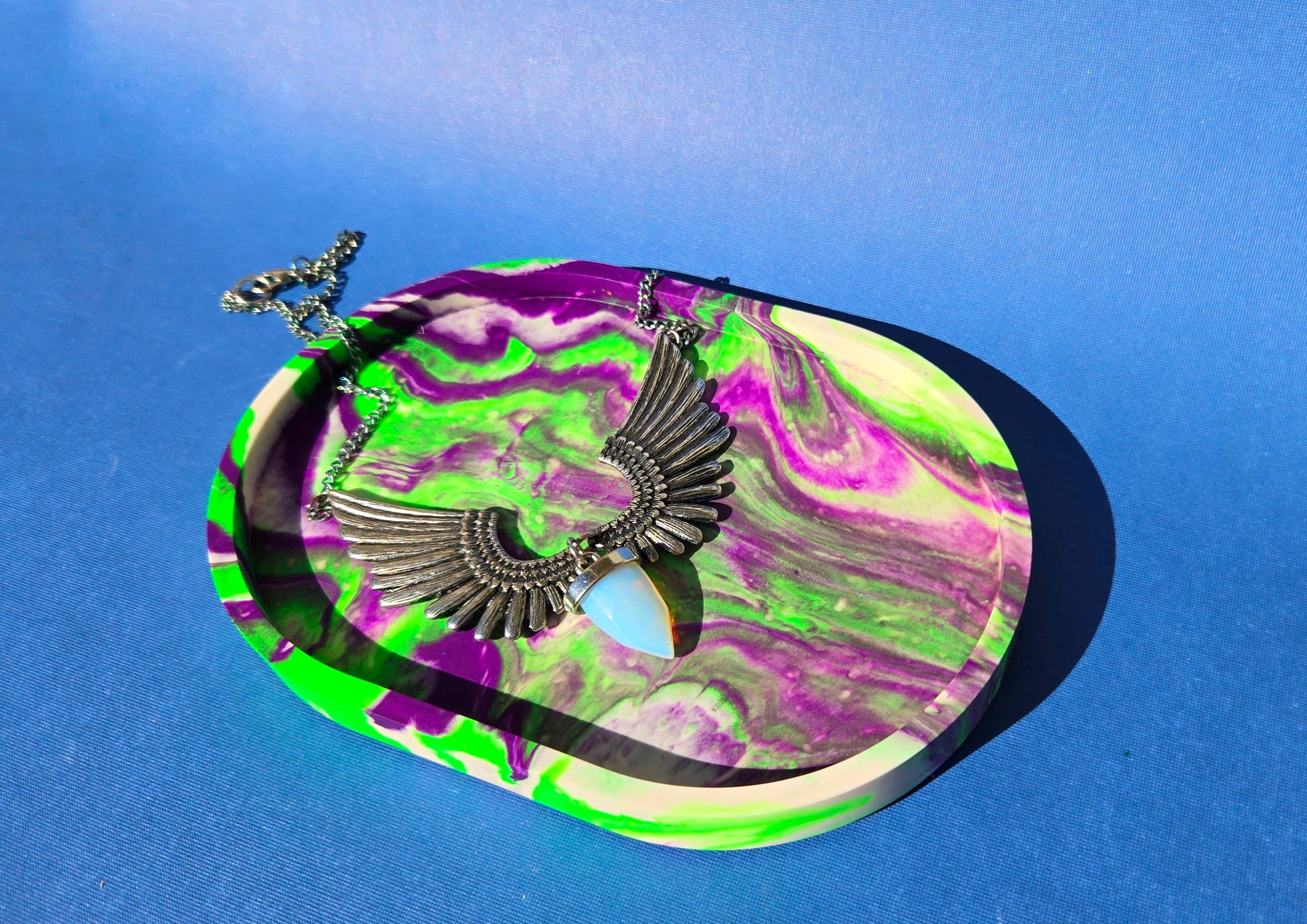 A jesmonite oval trinket tray or catch-all tray with bright green and purple swirls across the natural off-white surface. The tray is against a blue background with an opalite and wing pendant necklace resting in it.