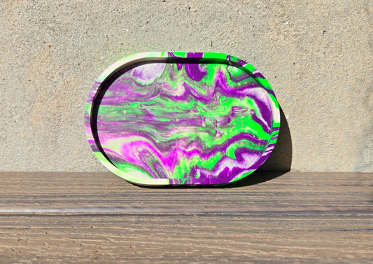 A jesmonite oval trinket tray or catch-all tray with bright green and purple swirls across the natural off-white surface. The tray is leaned up against a concrete wall and is resting on a wood plank.