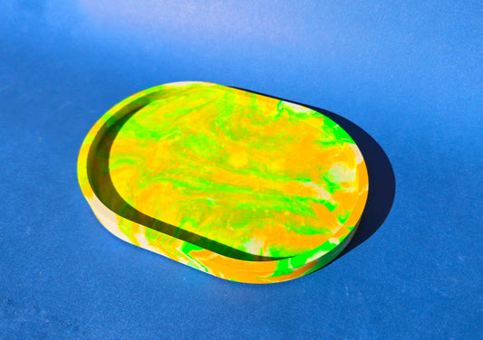 A jesmonite oval trinket tray or catch-all tray with bright green and yellow swirls across the natural off-white surface. The tray is against a blue background.