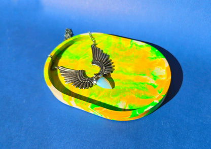 A jesmonite oval trinket tray or catch-all tray with bright green and yellow swirls across the natural off-white surface. The tray is against a blue background with an opalite and wing pendant necklace resting in it.