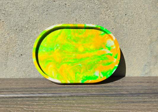 A jesmonite oval trinket tray or catch-all tray with bright green and yellow swirls across the natural off-white surface. The tray is leaned up against a concrete wall and is resting on a wood plank.