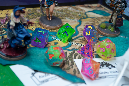 A 7-piece glitter dice set on top of a coastal map surrounded by 3 painted character miniatures. The dice are each 2 to 3 glitter colors, covering the whole rainbow across the entire set, with varying number colors to compliment each die.