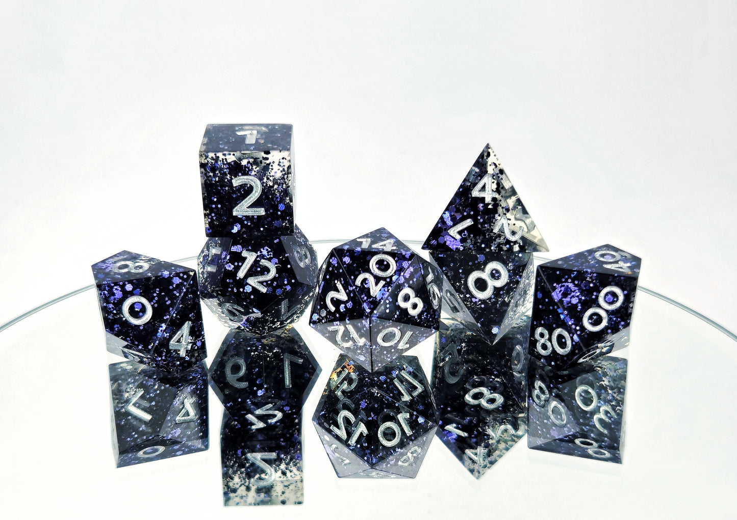 A 7 piece polyhedral ttrpg dice set. The dice are transparent clear with chunky dark navy blue glitter and bright silver numbers.