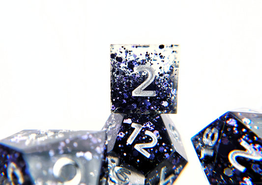 A closeup of a d6 stacked on top of a d12 from a ttrpg dice set. The dice are transparent clear with chunky dark navy blue glitter and bright silver numbers.