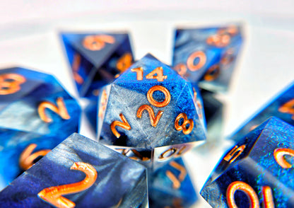 A 7 piece polyhedral ttrpg dice set. The dice are swirls of silver, shimmery navy, and shimmery bright blue with copper numbers.