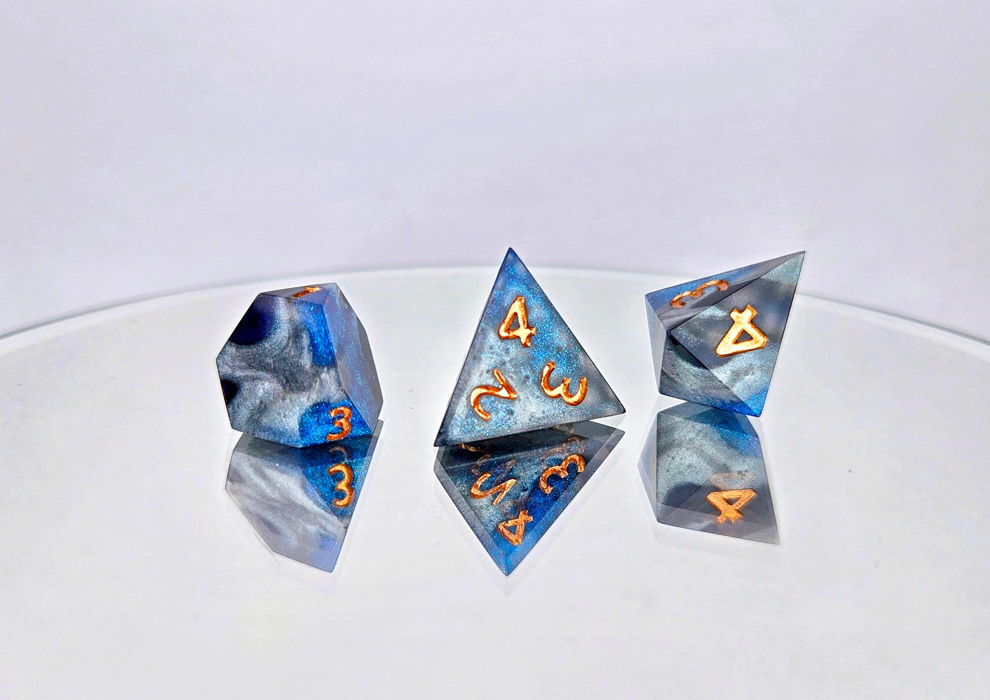 A comparison of 3 different d4 shapes for a ttrpg dice set. The shapes are a truncated d4, a standard caltrop d4, and a crystal shard style d4. The dice are swirls of silver, shimmery navy, and shimmery bright blue with copper numbers.