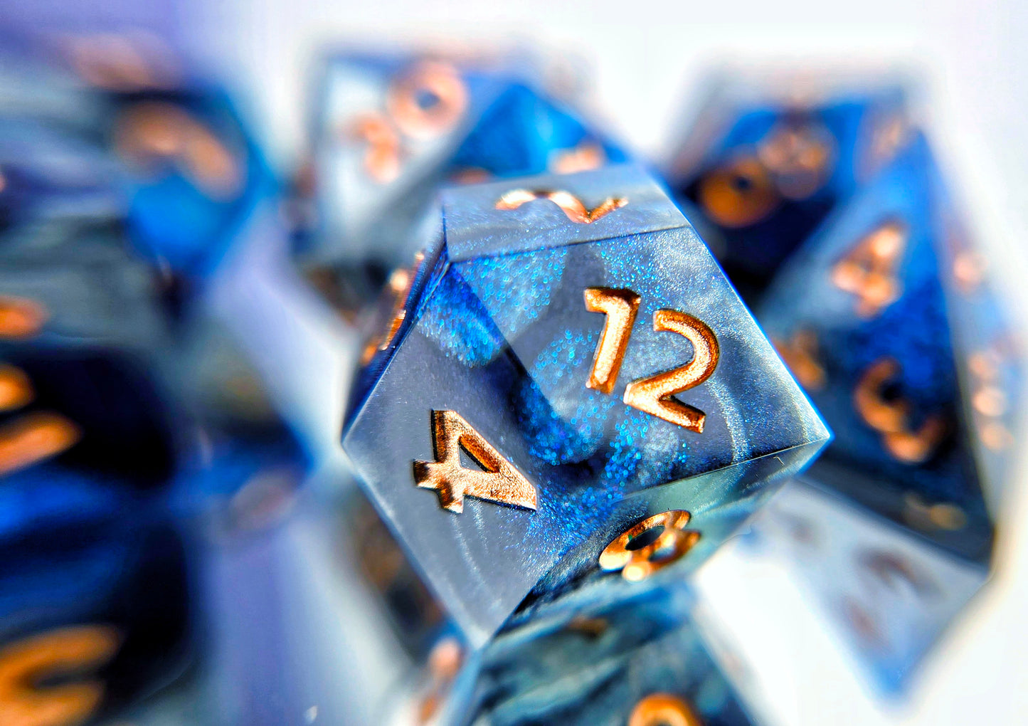 A closeup of a rhombic d12 from a ttrpg dice set. The dice are swirls of silver, shimmery navy, and shimmery bright blue with copper numbers.