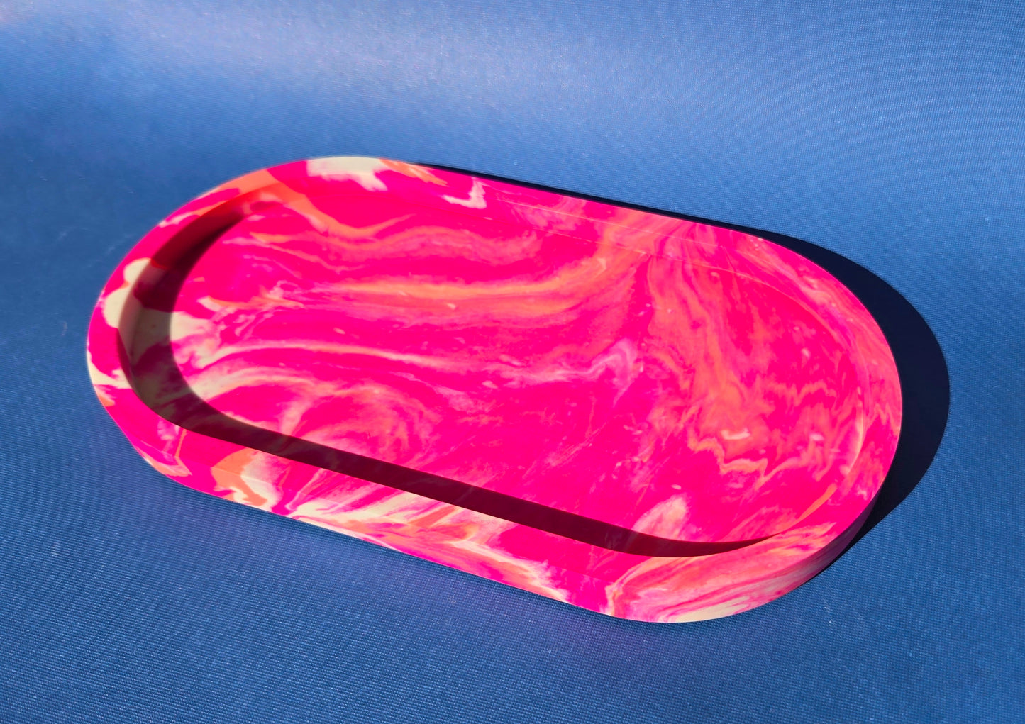 A jesmonite oval trinket tray or catch-all tray with bright pink and peachy orange swirls across the natural off-white surface. The tray is against a blue background.