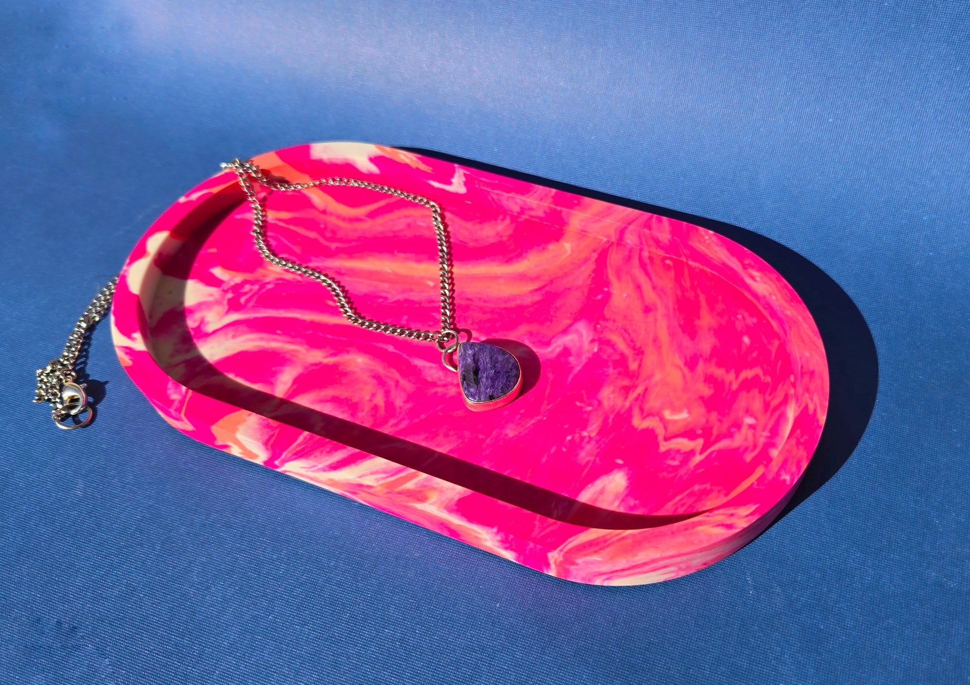 A jesmonite oval trinket tray or catch-all tray with bright pink and peachy orange swirls across the natural off-white surface. The tray is against a blue background and has a charoite necklace resting in it.