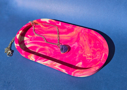 A jesmonite oval trinket tray or catch-all tray with bright pink and peachy orange swirls across the natural off-white surface. The tray is against a blue background and has a charoite necklace resting in it.