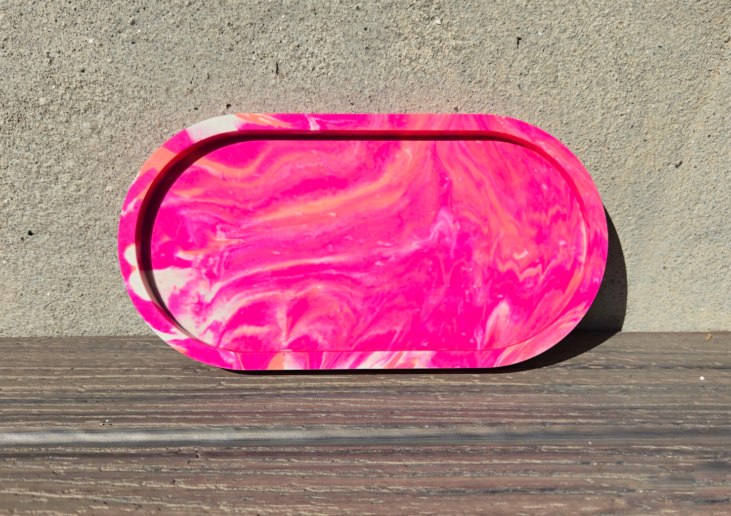 A jesmonite oval trinket tray or catch-all tray with bright pink and peachy orange swirls across the natural off-white surface. The tray is leaned up against a concrete wall and is resting on a wood plank.