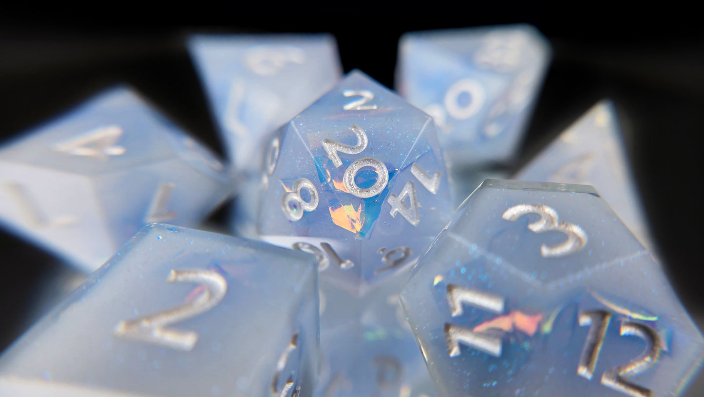 A 7 piece polyhedral ttrpg dice set. The dice are milky blue-ish white resin and blue/green sparkles with flashes of ice blue mylar peeking out and pearl white numbers.