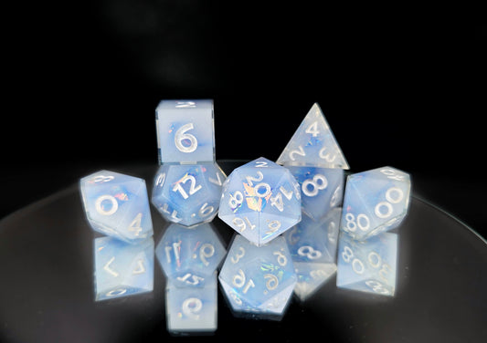 A 7 piece polyhedral ttrpg dice set. The dice are milky blue-ish white resin and blue/green sparkles with flashes of ice blue mylar peeking out and pearl white numbers.