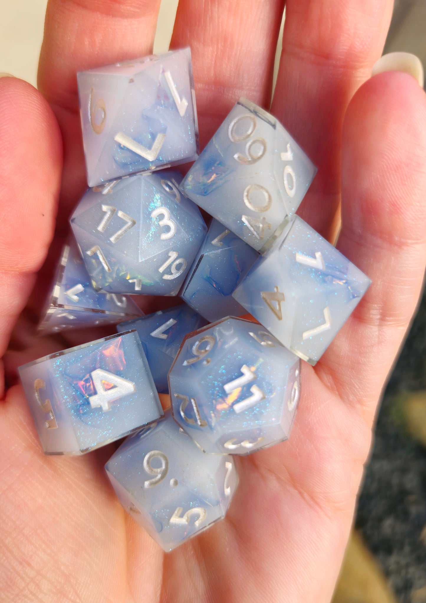 A polyhedral ttrpg dice set held in an open palm outside in the sunlight. The dice are milky blue-ish white resin and blue/green sparkles with flashes of ice blue mylar peeking out and pearl white numbers. 