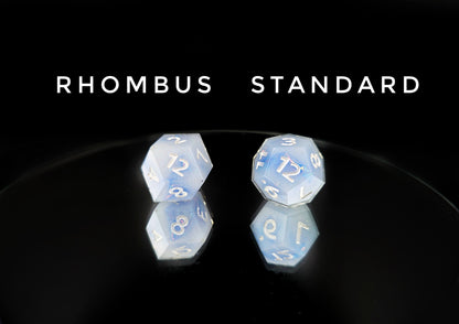 A comparison of different available d12 shapes from a ttrpg dice set. The dice are milky blue-ish white resin and blue/green sparkles with flashes of ice blue mylar peeking out and pearl white numbers. The text on the image reads "rhombus, standard", corresponding to the shapes photographed below them.