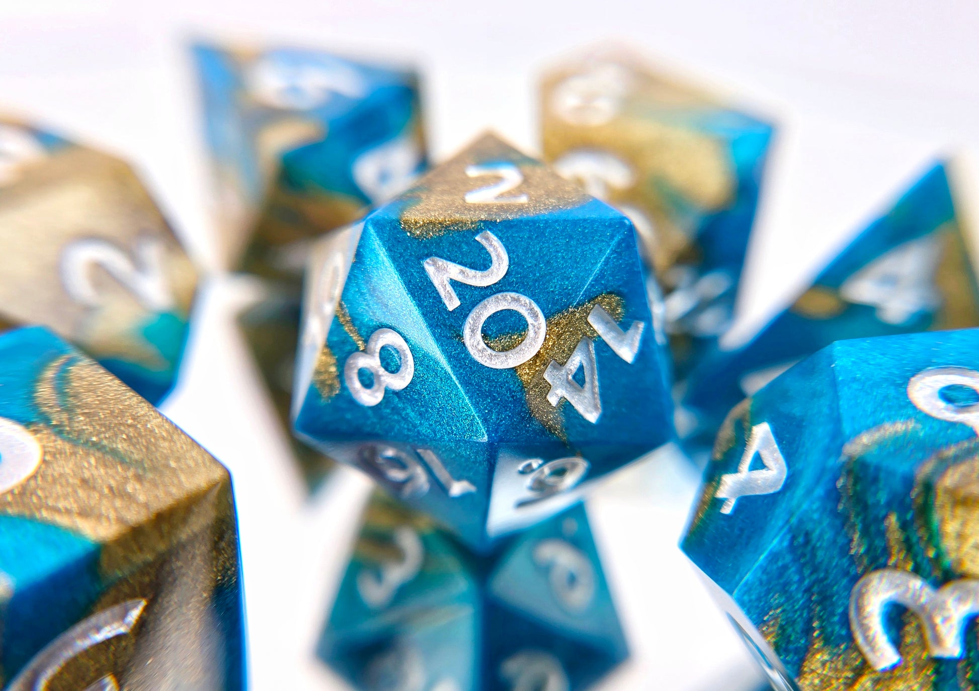 A 7 piece polyhedral ttrpg dice set. The dice are swirled metallic teal and gold with pearl white numbers.