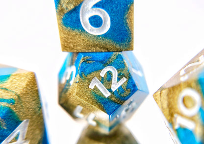 A closeup of a d12 from ttrpg dice set with a d6 stacked on top of it partially visible. The dice are swirled metallic teal and gold with pearl white numbers.