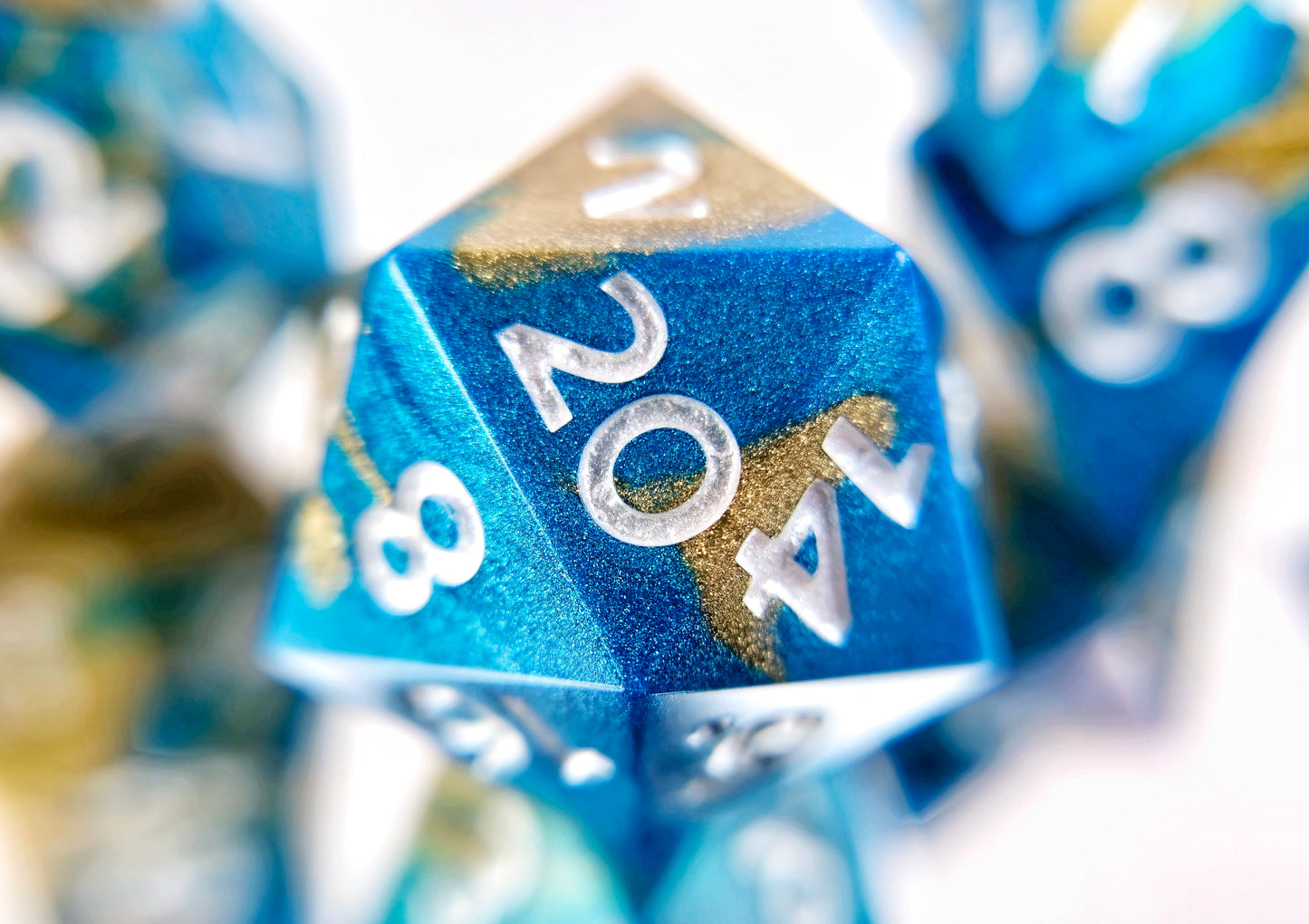 A closeup of a d20 from a ttrpg dice set. The dice are swirled metallic teal and gold with pearl white numbers.