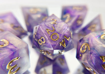 A 7 piece polyhedral ttrpg dice set. The dice are a warm pearlescent base with purple and navy ink swirled over top with soft gold numbers.