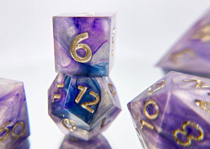 A closeup of a d6 stacked on top of a d12 from a ttrpg dice set. The dice are a warm pearlescent base with purple and navy ink swirled over top with soft gold numbers.