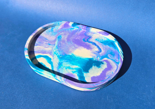 A jesmonite oval trinket tray or catch-all tray with teal blue and light purple swirls across the natural off-white surface. The tray is against a medium blue background.