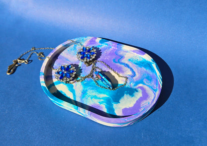 A jesmonite oval trinket tray or catch-all tray with teal blue and light purple swirls across the natural off-white surface. The tray is against a blue background and has a blue gemstone necklace resting in it.