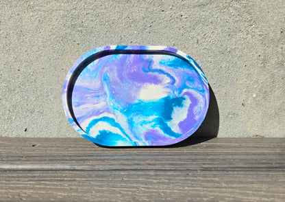 A jesmonite oval trinket tray or catch-all tray with teal blue and light purple swirls across the natural off-white surface. The tray is leaned up against a concrete wall and is resting on a wood plank.