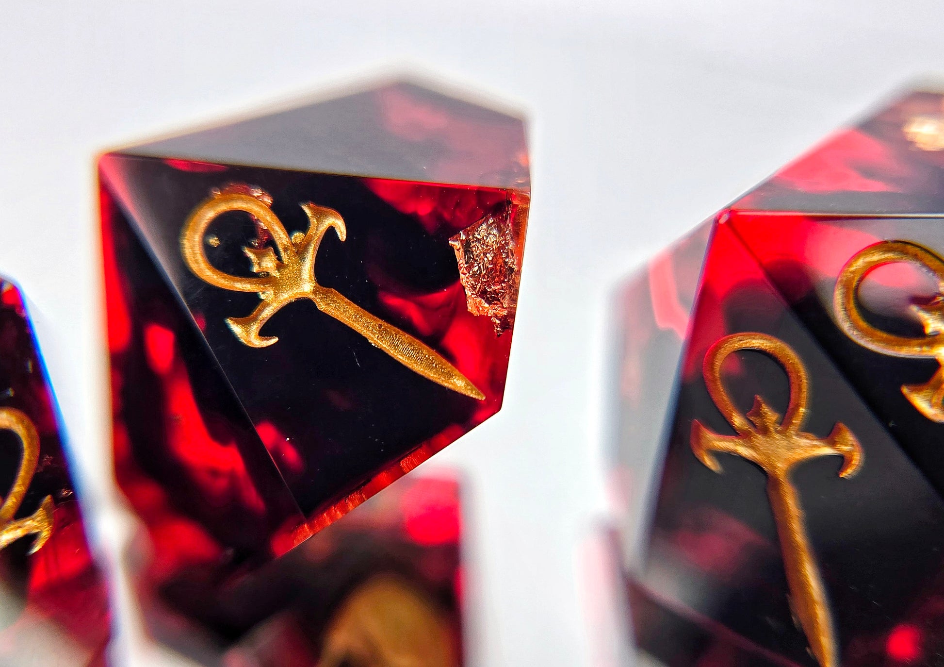 A closeup of a Vampire the Masquerade Hunger style die focusing on the standard ankh symbol. The dice are transparent red with black ink and gold foil inside. The symbols are inked in gold.