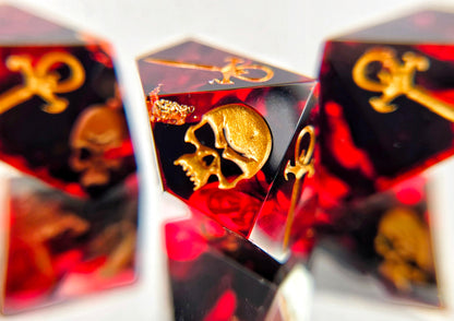 A closeup of a Vampire the Masquerade Hunger style die focusing on the skull. The dice are transparent red with black ink and gold foil inside. The symbols are inked in gold.