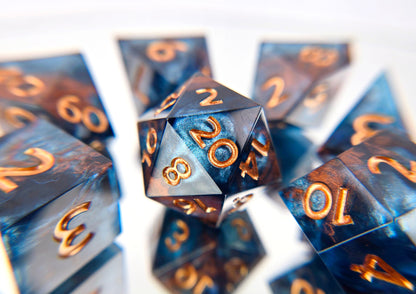 A full 7 piece polyhedral ttrpg dice set. The dice are transparent dark blue with bloops of shimmery silver inside and streaks of bronze coloring across the surface and warm copper numbering. 