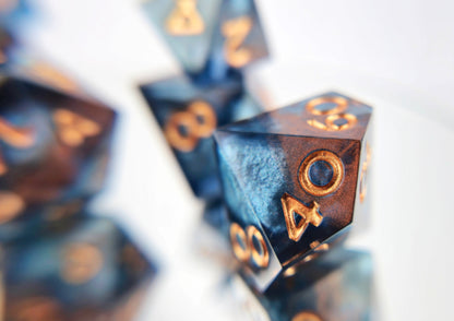 A closeup of a d100 from a ttrpg dice set. The dice are transparent dark blue with bloops of shimmery silver inside and streaks of bronze coloring across the surface and warm copper numbering. 