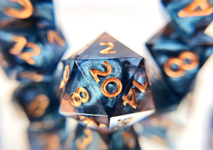 A closeup of a d20 from a ttrpg dice set. The dice are transparent dark blue with bloops of shimmery silver inside and streaks of bronze coloring across the surface and warm copper numbering. 