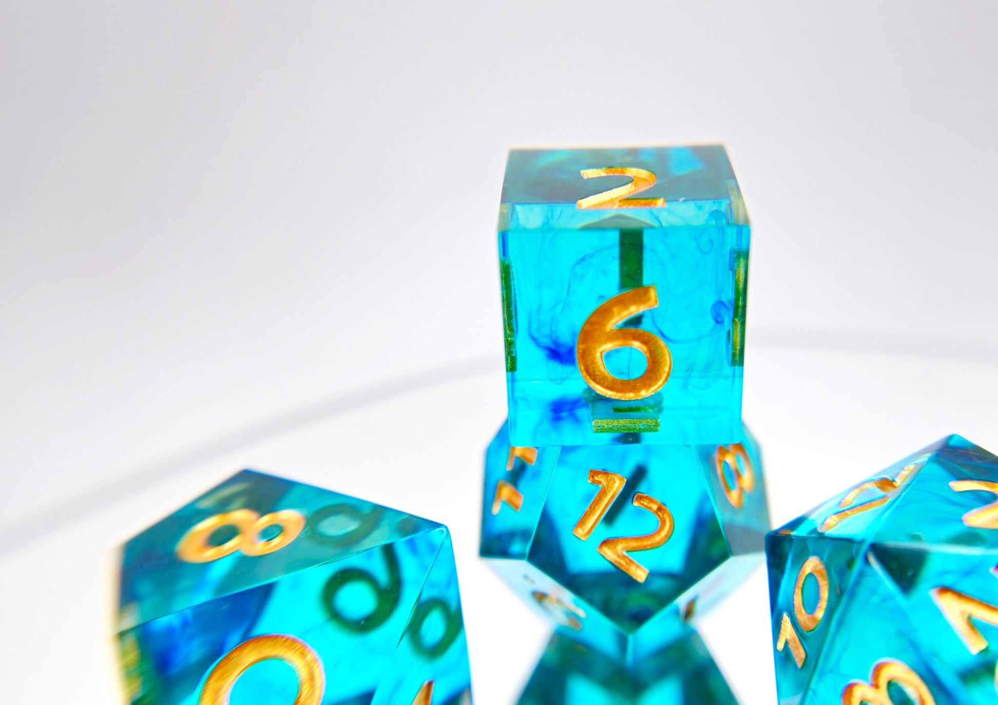 A closeup of a d6 sitting on top of a d12 from a polyhedral ttrpg dice set. The dice are transparent light teal blue with thin wisps of darker blue swirled inside and gold numbering. 