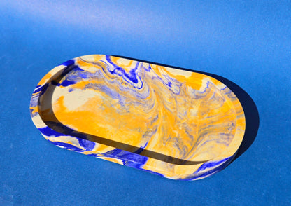 A jesmonite oval trinket tray or catch-all tray with blue and tan swirls across the natural off-white surface. The tray is against a blue background.