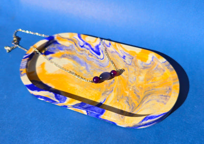 A jesmonite oval trinket tray or catch-all tray with blue and tan swirls across the natural off-white surface. The tray is against a blue background and has a necklace with purple beads resting in it.