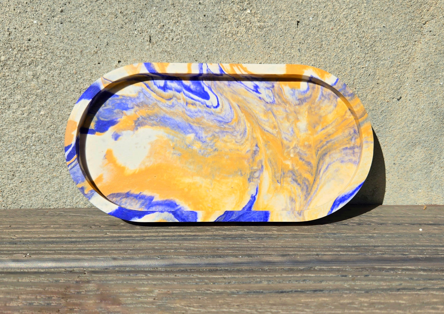 A jesmonite oval trinket tray or catch-all tray with blue and tan swirls across the natural off-white surface. The tray is leaned up against a concrete wall and is resting on a wood plank.