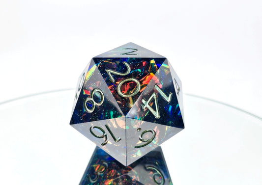 An oversized d20 on a mirror and against a white background. The die is a transparent dark blue resin with super fine green glitter sparkles and intense red to orange to yellow to green colorshift mylar foil crinkles inside. The numbers are inked a greenish bronze.